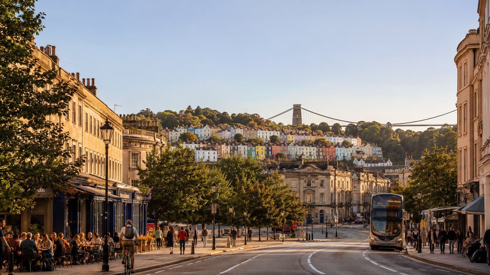 A lively Bristol street at golden hour with cafés, cyclists and a city bus, the Clifton Suspension Bridge beyond